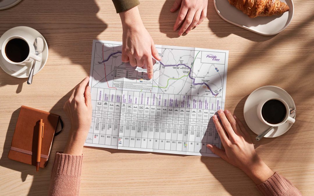 A Purple Line MD - LRT Station Features Map sits on a table. Hands of people reviewing the map, pointing to specific locations. Coffee mugs surround the map.